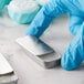 A hand in blue gloves holding a silver metal mini tin with a slide top.