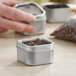 A hand holding a silver square tin with a clear window lid containing herbs.