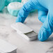 A hand in a blue glove holding a small white rectangular tin with a slide top.