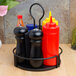 A Tablecraft black metal condiment caddy on a table with three condiments.