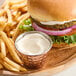A small metal cup filled with creamy vegan mayonnaise is placed next to French fries and a veggie burger on a wooden plate.