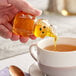 A hand holding a small clear plastic bear pouring honey into a cup of tea.