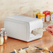 A person putting a white napkin into a white rectangular countertop napkin dispenser.