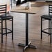 A square bar height table with a light wood laminate top and a black cast iron base.