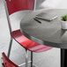 A round bar height table with a gray laminated top, accompanied by red chairs, a small potted plant, a notebook, and a pen on the tabletop.