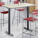 A rectangular bar height table with a reversible gray and white laminated top and a cast iron base, surrounded by red and metal bar stools.
