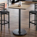 A square bar height table with a light wood laminate top and a black cast iron pedestal base.