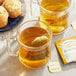 A glass mug of Twinings Buttermint herbal tea with a tea bag in it on a table with muffins.