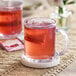 A clear glass mug filled with reddish herbal tea and a tea bag, placed on a white coaster.