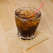 A glass with a straw and brown liquid with a beige Hoffmaster cocktail napkin on the table.