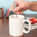 A hand holding a spoon in a white Tuxton china mug.