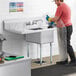 A man in a plastic apron and gloves cleaning a Regency commercial sink.