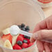 A hand using a spoon to add Add A Scoop Vitamin C Blend supplement powder to a bowl of fruit.