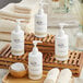 A white bottle of Novo Essentials Hotel and Motel Body Wash with a white cap on a wooden tray with white towels and a bowl of cotton pads.