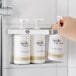 A hand holding a white plastic pump dispenser with Novo by Noble Chemical Triple Bottle Bracket for hotel amenities on a shelf.