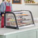 An Avantco curved refrigerated countertop bakery display case on a counter with food in it and a man wearing a red apron standing behind it.