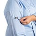 A woman wearing a sky blue Uncommon Chef Vigor Pro Vent chef coat with a mesh back putting a cell phone in the pocket.