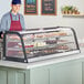 An Avantco curved black countertop bakery display case filled with pastries and cupcakes with a man standing behind it.