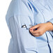 A person putting a cell phone in the pocket of a blue Uncommon Chef Vigor Pro Vent long sleeve chef coat with a mesh back.