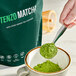 A hand holding a spoonful of Tenzo Premium Matcha Green Tea Powder.