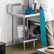 A man in a white shirt and blue apron standing next to a Regency stainless steel dish table undershelf.