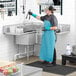 A man in a blue apron and gloves standing at a Regency stainless steel 3 compartment sink.