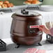 A person using an Avantco copper soup kettle warmer to heat soup.