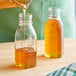 A person pouring honey from a glass bottle into a small glass Muth jar.