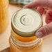 A hand holding a 70/450 gold metal lid over a jar of yellow liquid.