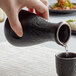 A hand pouring liquid from a black Acopa Heika sake bottle into a small black cup.