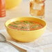 A Citrus Yellow Acopa Capri Bistro Bowl filled with soup with a spoon next to it.