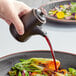 A hand pouring soy sauce from a black stoneware pourer onto a bowl of vegetables.