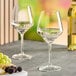 Two Acopa Silhouette wine glasses filled with white wine on a table with grapes and cheese.