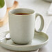 A white Acopa Pangea porcelain cup of coffee on a saucer with a spoon on a table.