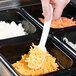 A person holding a white Cambro salad bar spoon over food.