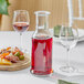 A Stolzle glass carafe filled with red liquid on a table next to a plate of fruit and wine.