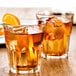 Two Fortessa Basics Chez Bistro Rocks glasses of brown liquid with ice and orange slices on a table.