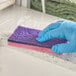 A gloved hand using a Lavex pink and purple sponge to clean a counter.