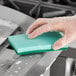 A hand in a plastic glove using a green Lavex sponge to clean a stainless steel sink.