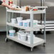 A person using a Rubbermaid three shelf utility cart to hold plastic containers of food.