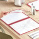 A woman's hand holding a burgundy 6-view trifold menu cover on a table.