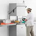 A man in a white coat and gloves using a Main Street Equipment door-type dish machine to wash dishes on a counter in a professional kitchen.