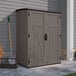 A brown Suncast vertical storage shed next to a wall with a shovel and rake inside.