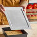 A rectangular metal deep dish pizza pan separator or lid being held above a pizza pan.