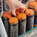 A hand holding a PakTech Tropical Orange plastic can carrier with a black beverage can inside.