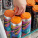 A hand opening a PakTech Tropical Orange plastic can carrier with 4 colorful cans inside.