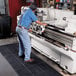 A man in a blue shirt and jeans standing on a black Cactus Mat Connectable Anti-Fatigue Floor Mat while working on a machine.