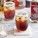 A bottle of Runamok Coffee-Infused Maple Syrup on a table with a glass of brown liquid and ice with a candle.