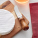 An Acopa stainless steel cheese spreader with a dark wood handle on a cutting board next to a round white cheese wheel.