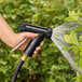 A hand holding a black Sani-Lav spray nozzle attached to a hose spraying water on a bush.